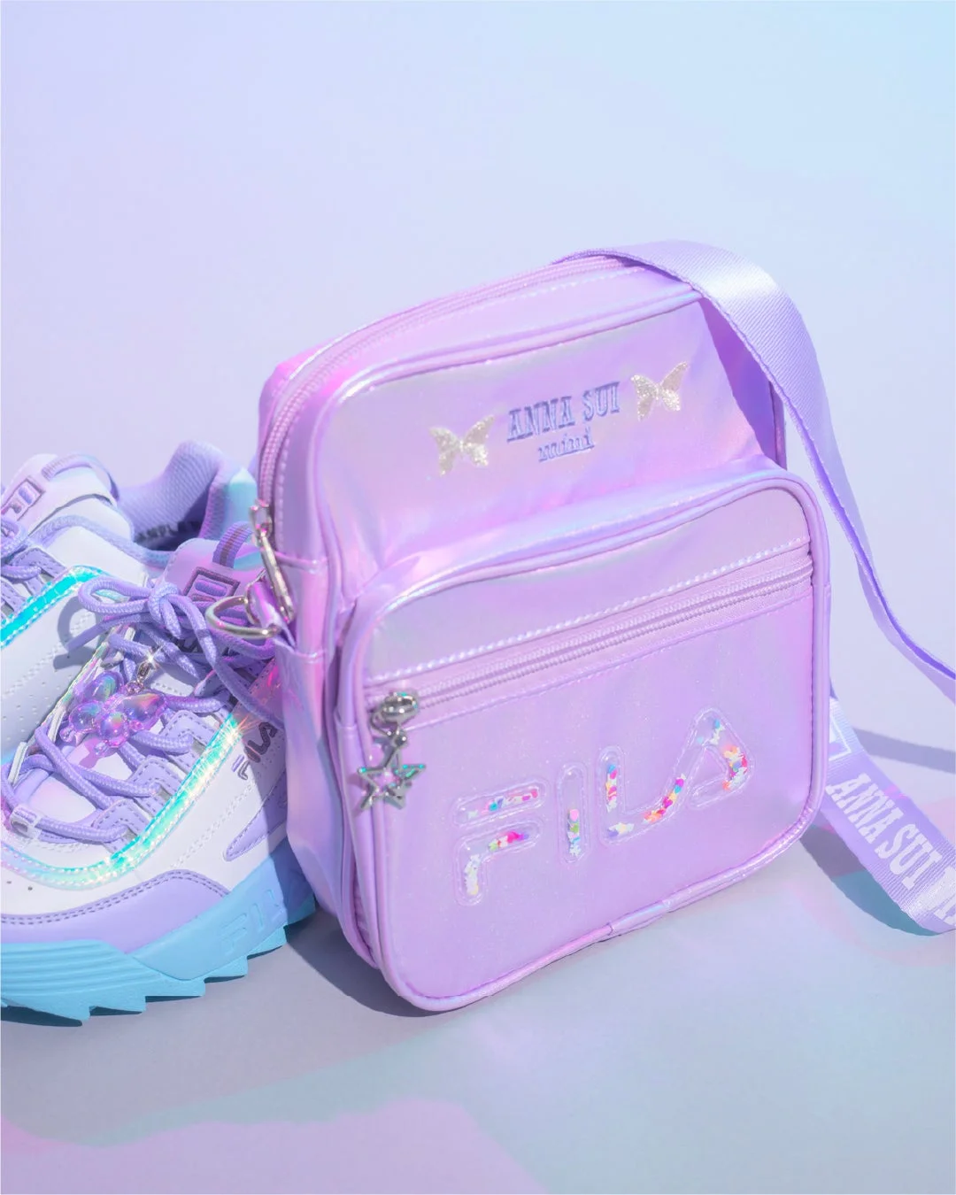 紫色の光沢あるショルダーバッグとスニーカーが並べられています。バッグには「ANNA SUI mini」と「FILA」のロゴ、蝶のモチーフがあり、全体的にパステルカラーとキラキラした質感が特徴的で、可愛らしいファッションアイテムの組み合わせです。