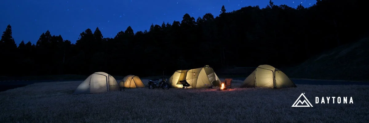 夜の広々とした野原で、いくつかのテントが明かりを灯し、焚き火が燃えている。奥には星が輝く夜空と暗い森が広がり、手前にはバイクも見える、静かで美しいキャンプの風景