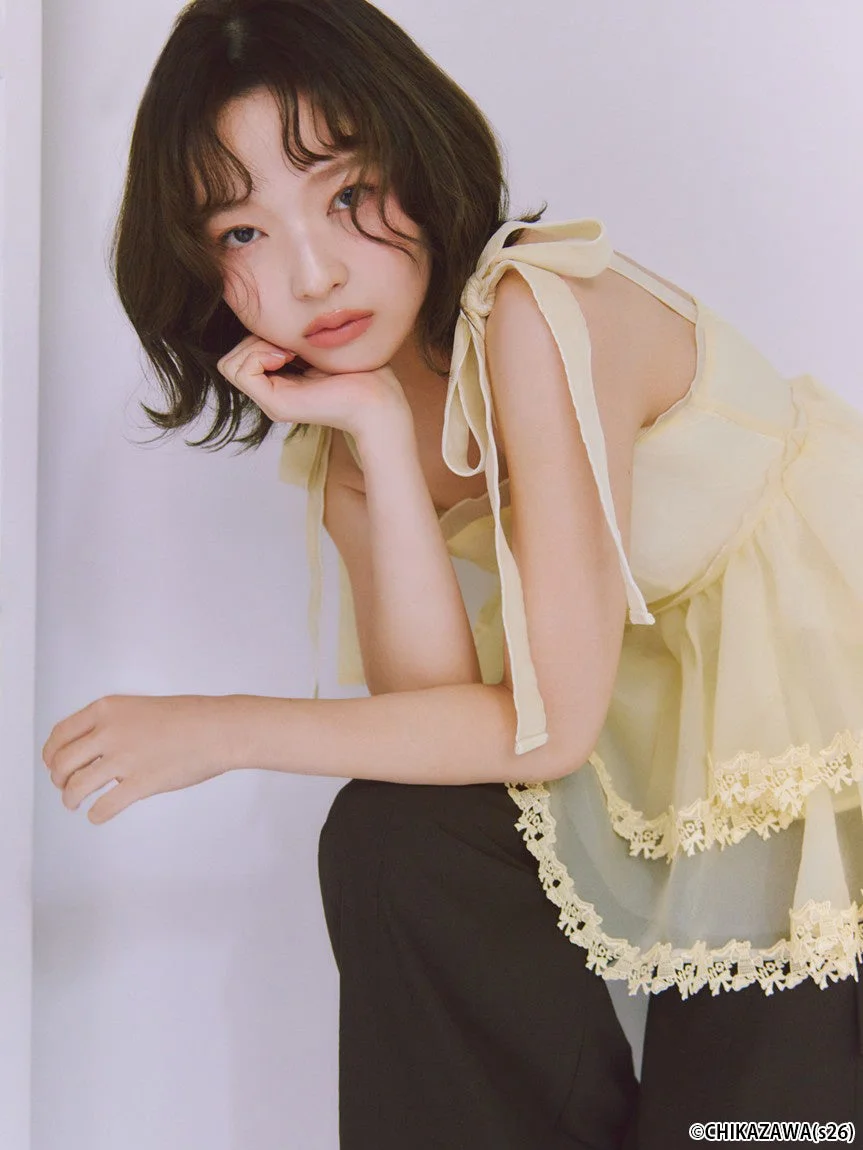 淡い黄色のレース付きトップスと黒いパンツを着用したショートヘアの女性が、肘をついてカメラを見つめているポートレート写真です。