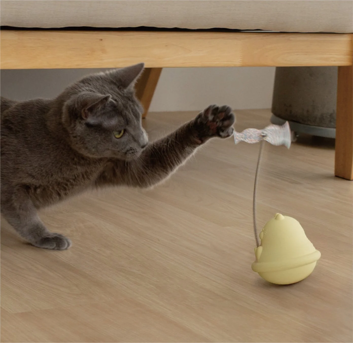 灰色の猫が、室内で黄色のタンブラー型のおもちゃで遊んでいる様子