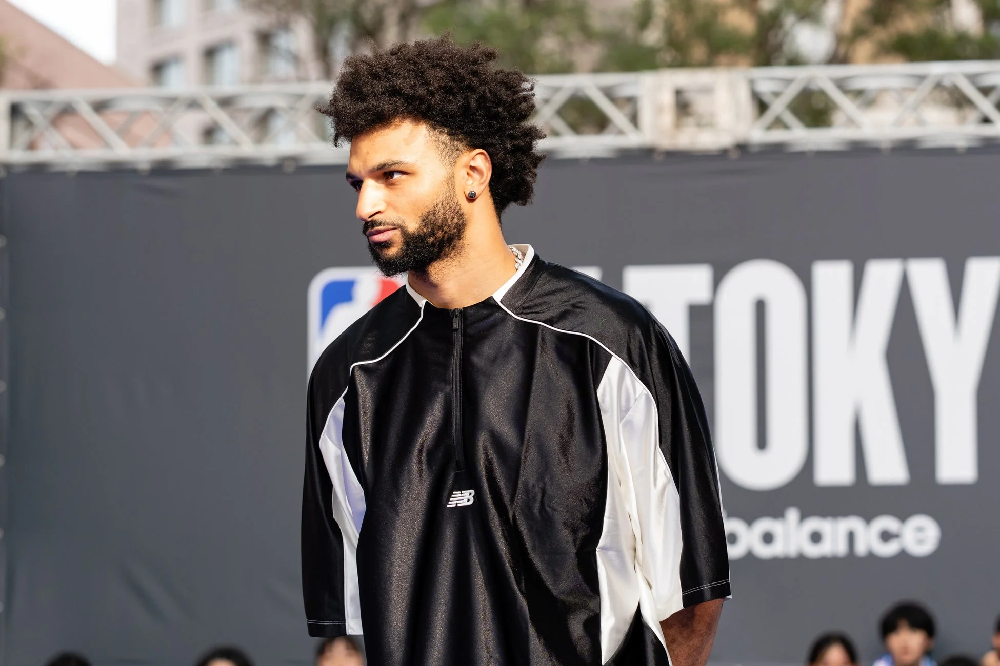 アフロヘアと髭が特徴的な男性が、黒と白のニューバランスのシャツを着て屋外イベントに立っている様子。