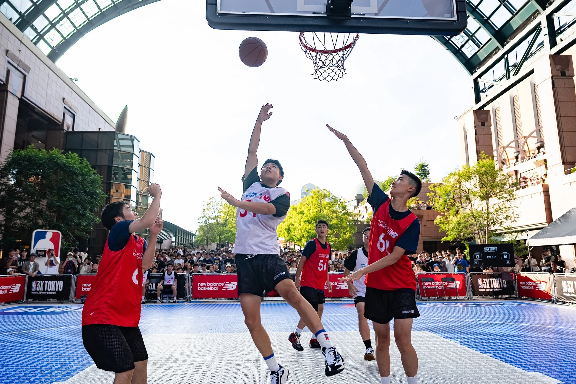 屋外の特設コートで3x3バスケットボールの試合が行われています。選手たちはニューバランスのユニフォームを着用し、一人がシュートを放とうとしている瞬間です。