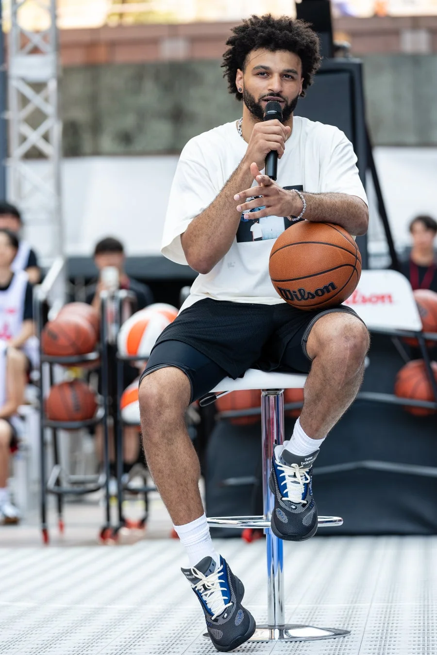 巻き毛の男性が屋外のバスケットボールイベントで、マイクを手に座って話しています。