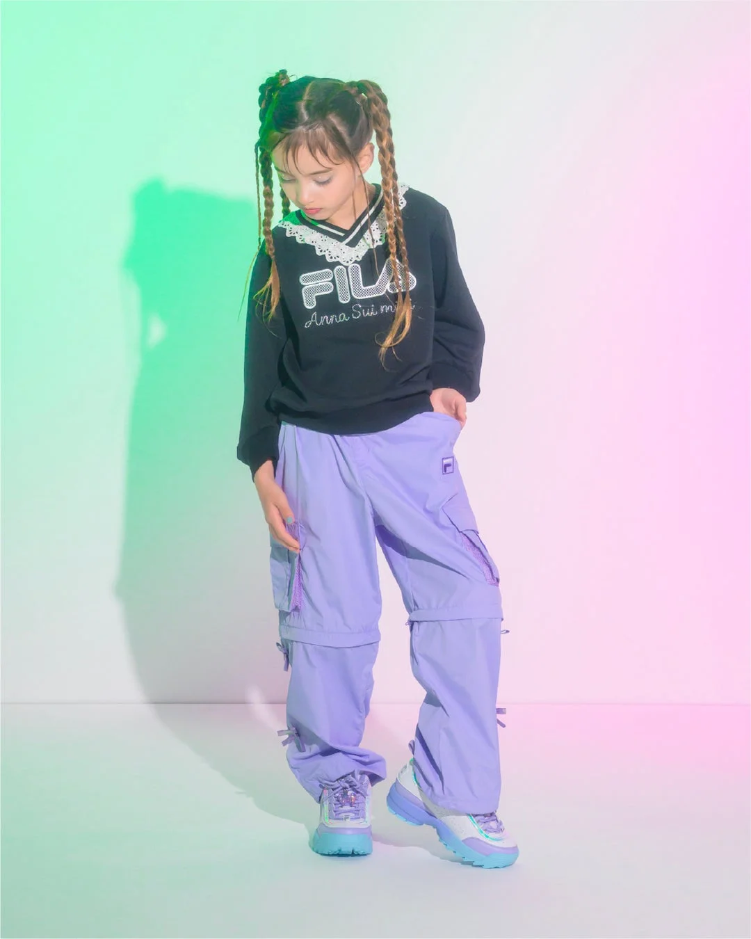 編み込みヘアの女の子が、黒いFILAとAnna Sui miniのコラボスウェットに紫のカーゴパンツ、カラフルなスニーカーを合わせたストリート系ファッションを着こなしています。ポップな背景が特徴的です。