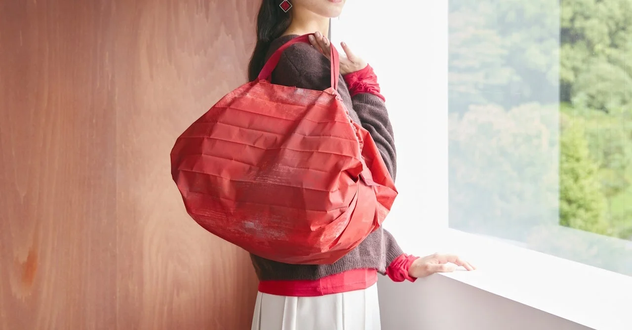 大きな赤いプリーツバッグを持った女性が、屋内の窓辺に立っています。茶色のセーターと白いスカートを着用しており、窓の外には緑豊かな木々が見える穏やかな雰囲気の画像です。