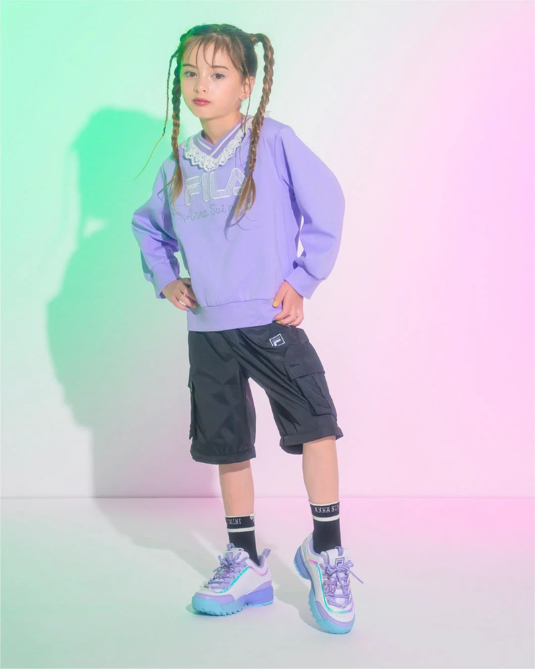 ツインテールの女の子が、パープルのスウェットと黒のショートパンツ、カラフルなスニーカーを着用し、両手を腰に当ててポーズをとっています。背景は緑とピンクのグラデーションで、明るくポップな雰囲気のキッズファッション写真です。