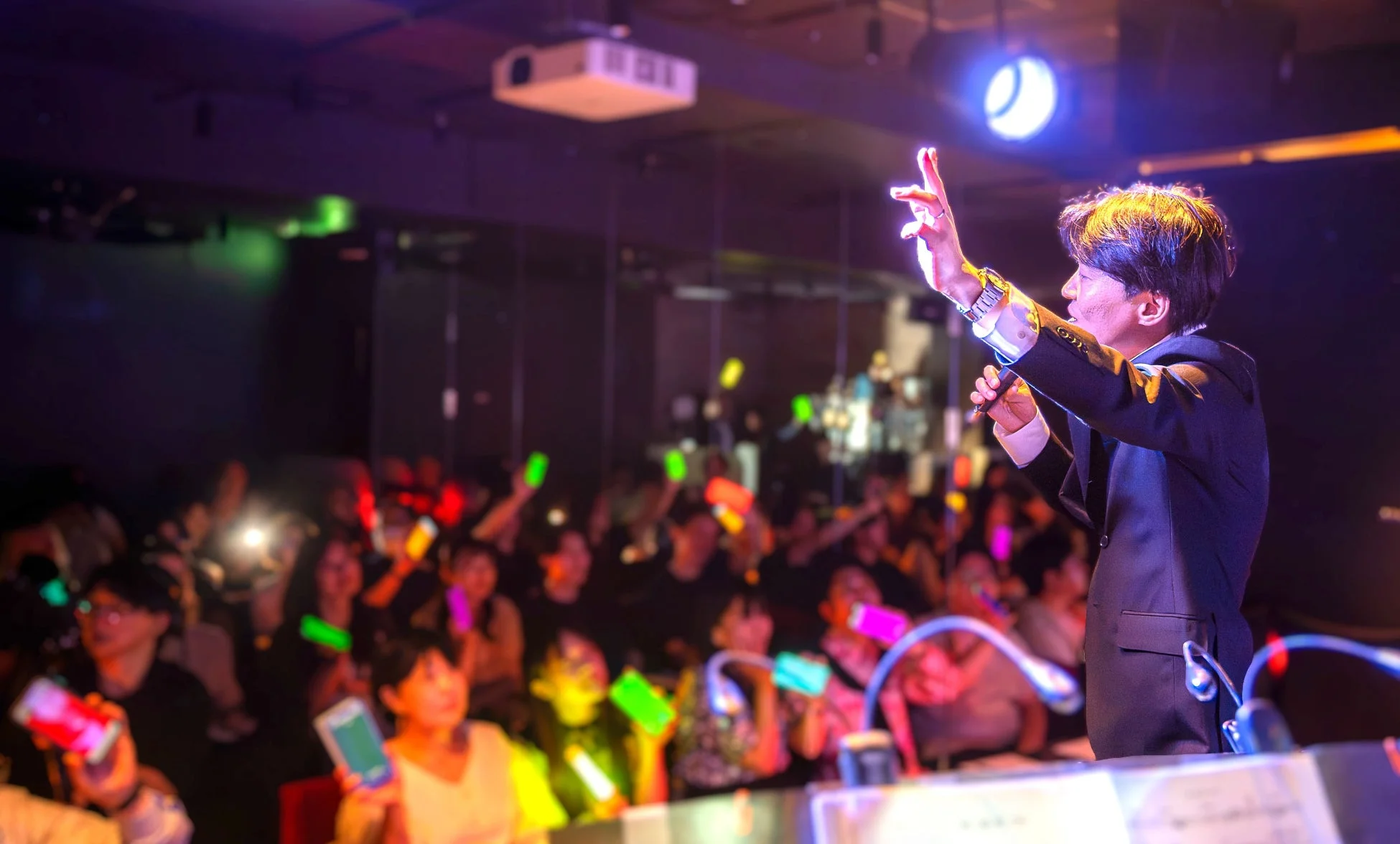 ライブイベントで、ステージ上の男性がマイクを持ち、観客に向かってパフォーマンスをしています。観客は色とりどりのペンライトやスマートフォンを掲げ、熱気あふれる雰囲気が伝わってきます。