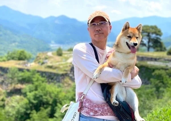 男性が柴犬を抱っこしてカメラに微笑みかけている。背景には美しい山々が広がる自然豊かな風景が写っている。