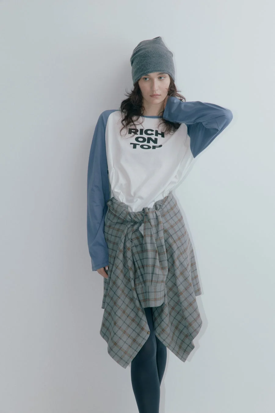 白い背景の前に立つカジュアルな服装の若い女性の全身像です。グレーのビーニー帽、白と青のラグランスリーブTシャツ、腰に巻いたチェック柄のシャツ、黒いレギンスを着用しています。