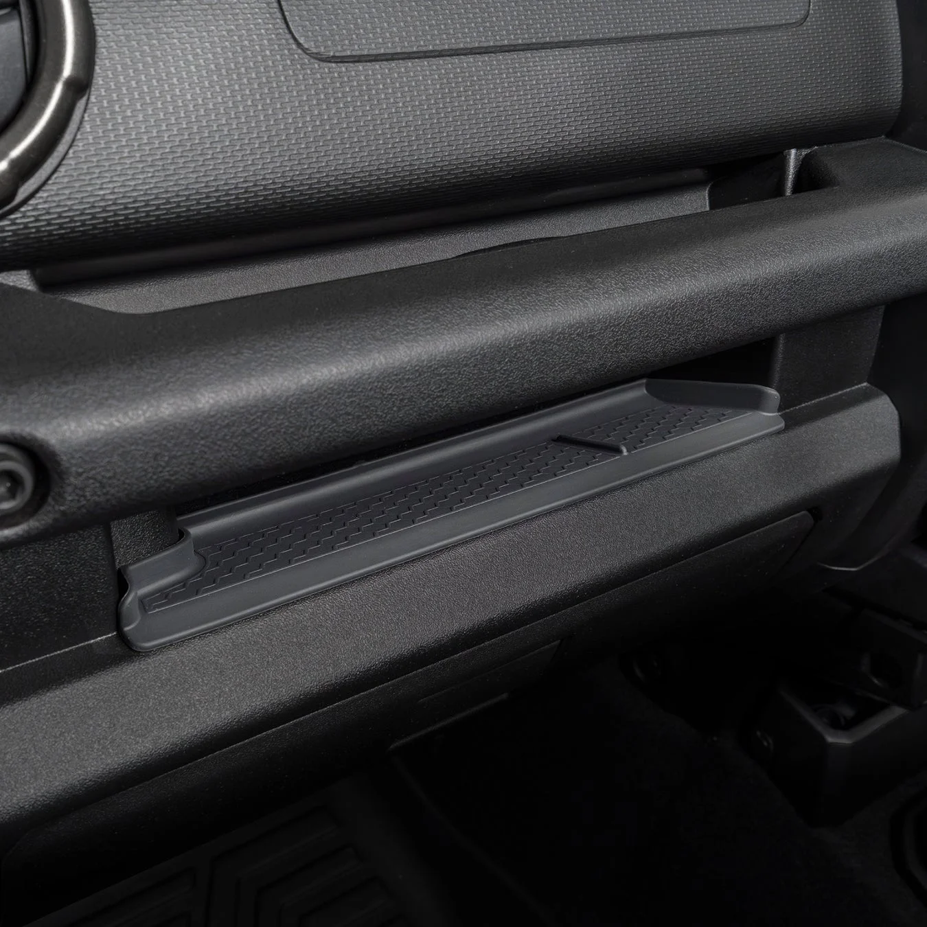 自動車の助手席側ダッシュボード周辺をクローズアップした画像です。黒いプラスチック製の内装には、グローブボックス上部に横長の収納トレイと、その上部に頑丈な取っ手が見えます。表面には細かい凹凸加工が施されています。