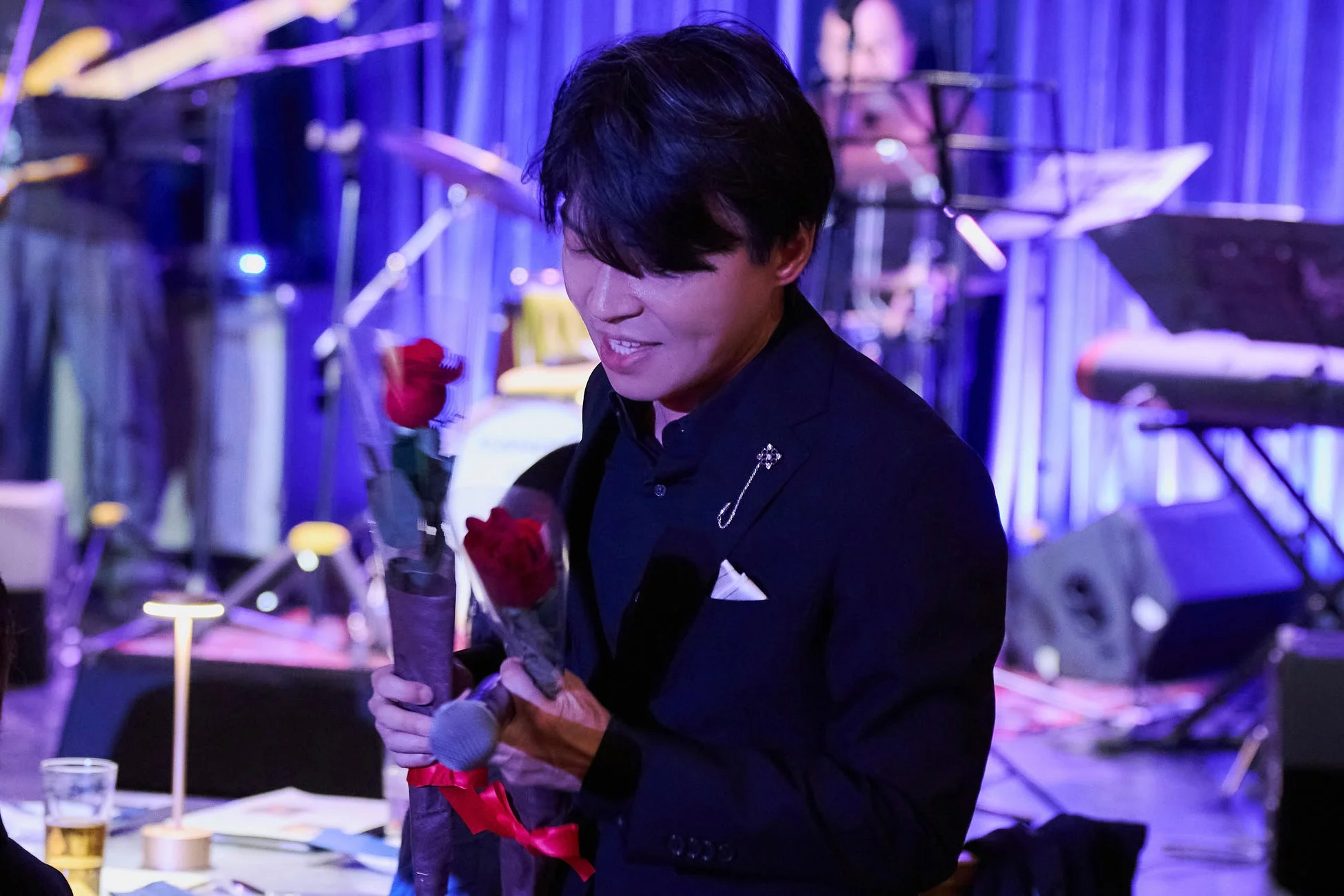 青い照明に照らされたステージで、スーツを着た男性が赤いバラの花束とマイクを持っている様子です。