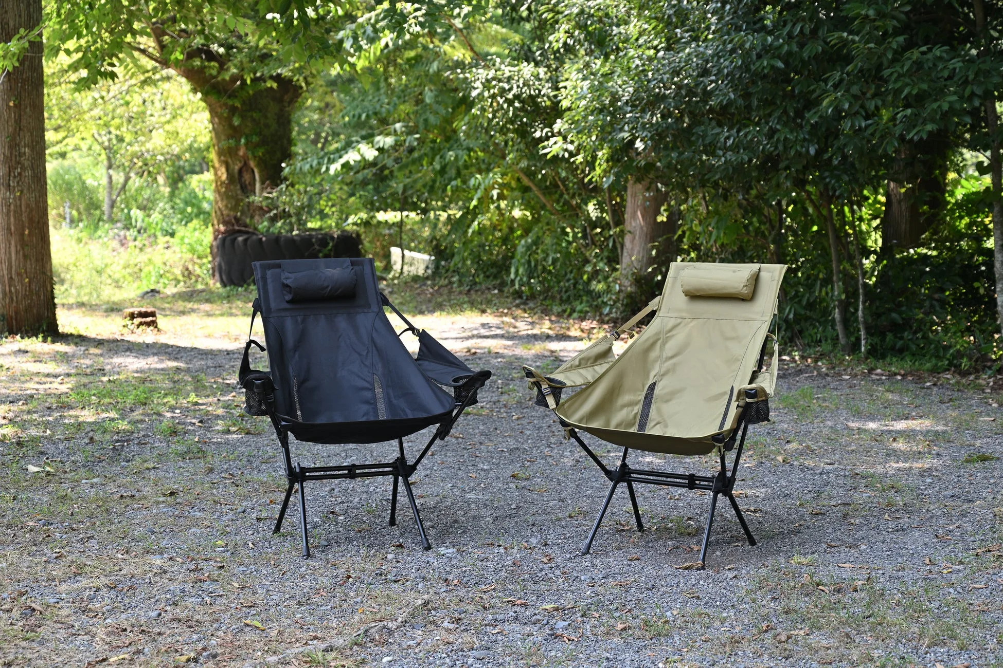 木々の茂る自然の中で、黒とカーキ色のアウトドアチェアが2脚、砂利の地面に置かれている