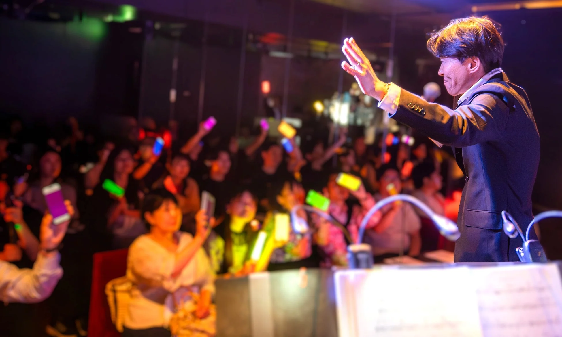 ライブステージで歌唱または指揮をする男性と、カラフルな光るスマートフォンやペンライトを掲げて盛り上がる観客の様子。熱気あふれるイベントの瞬間を捉えています。