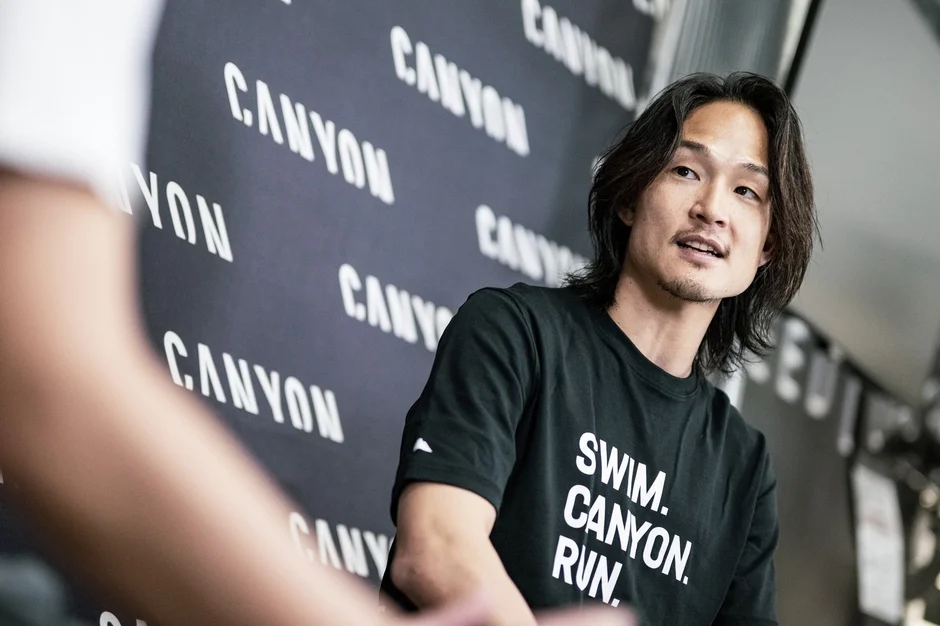 「CANYON」のロゴが並ぶ壁を背景に、黒いTシャツを着たアジア人男性が話している。