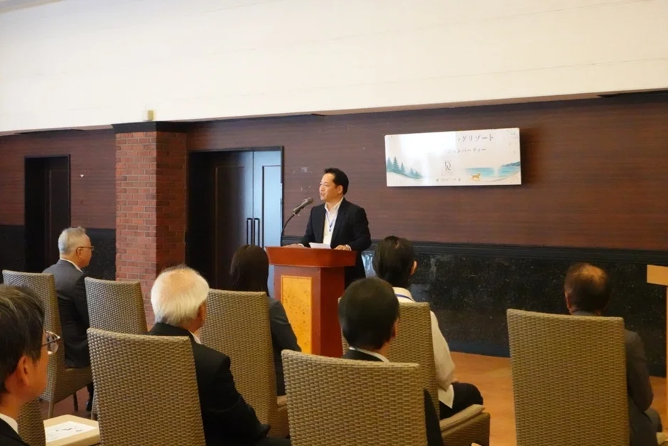 室内で男性が壇上でスピーチをしており、複数の聴衆が着席して聞いています。