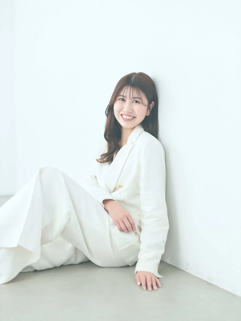 白いスーツを着た女性が、白い壁に寄りかかり床に座って微笑んでいます