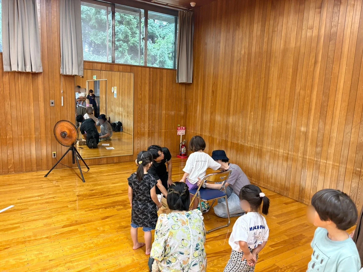 木目調の壁と床の広い室内で、数人の子供と大人が集まって活動している様子です。