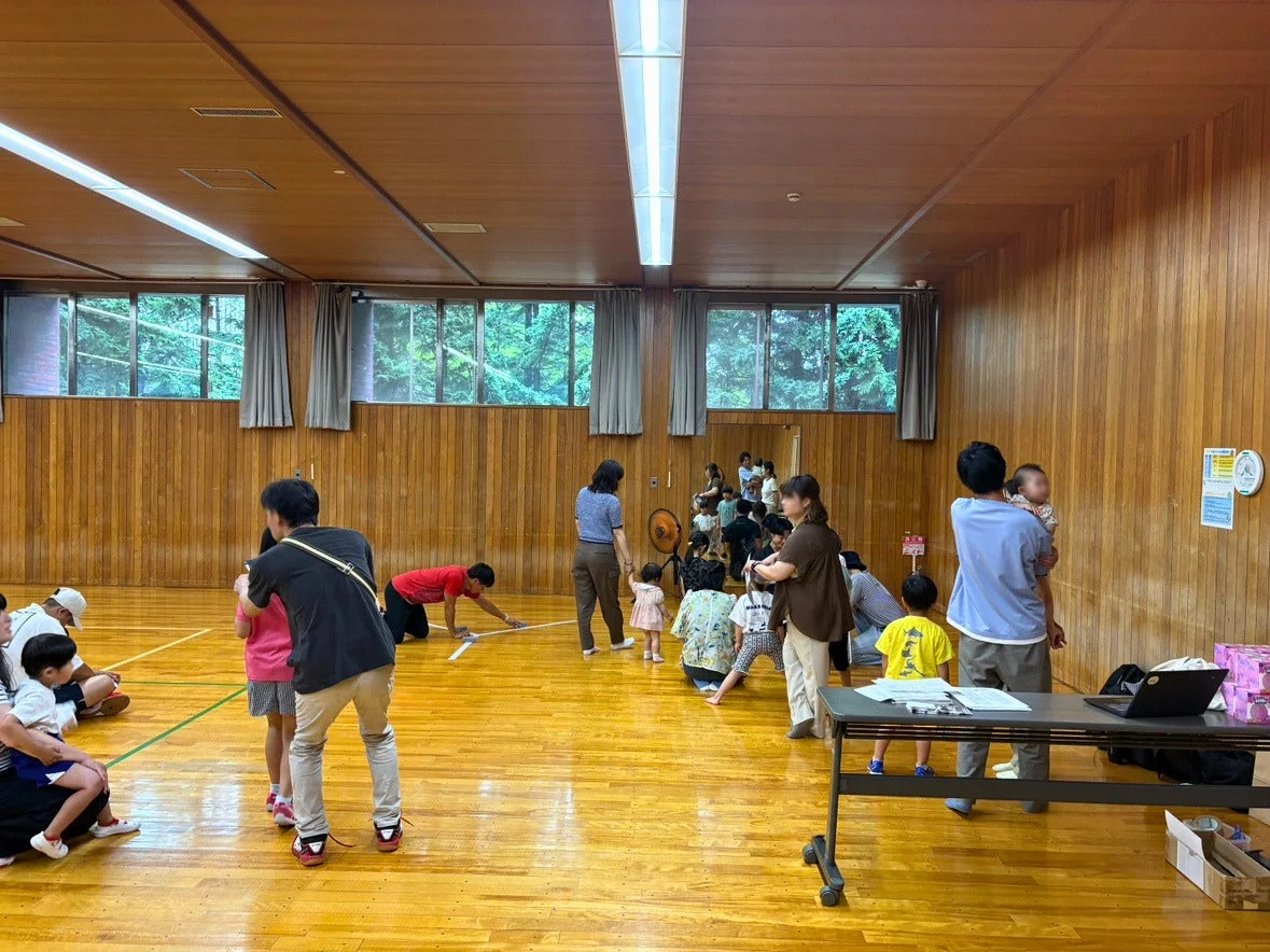 体育館のような広間で、大人と子供たちが集まり、何らかの活動やイベントに参加している様子が写っています。