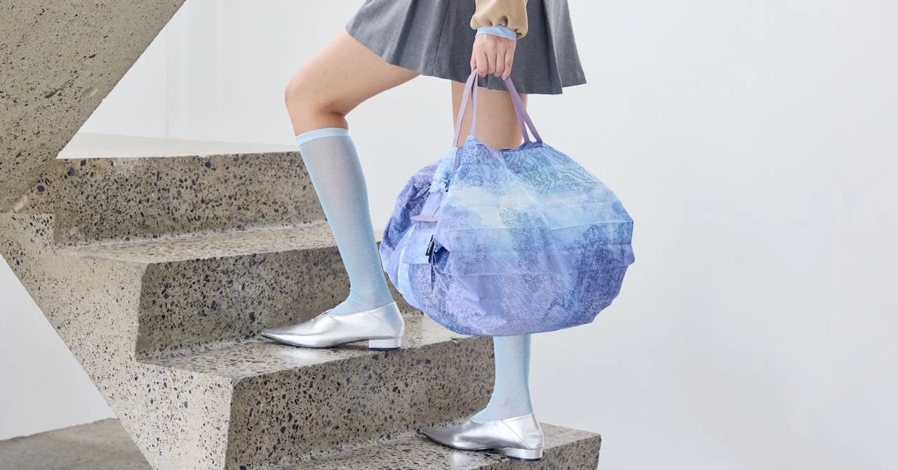 階段を上る女性が、プリーツスカートに水色の靴下と銀色の靴を合わせ、水色と紫のグラデーションが特徴的な大きなトートバッグを提げています。