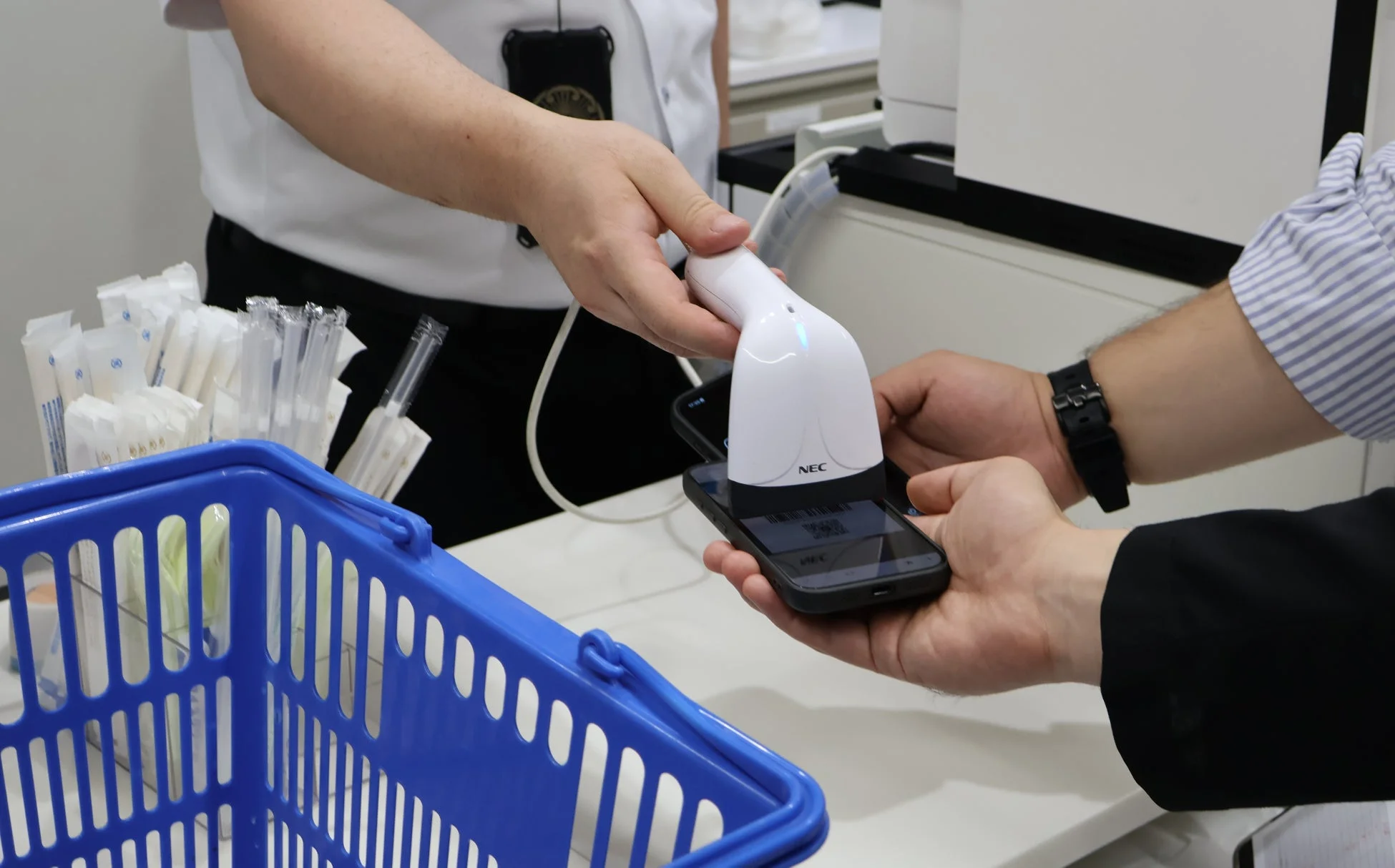 店員がNECのバーコードスキャナーを使って、顧客のスマートフォンに表示されたQRコードを読み取っている様子です。支払いかサービス受付の場面で、近くには青いカゴに入った商品が見えます。