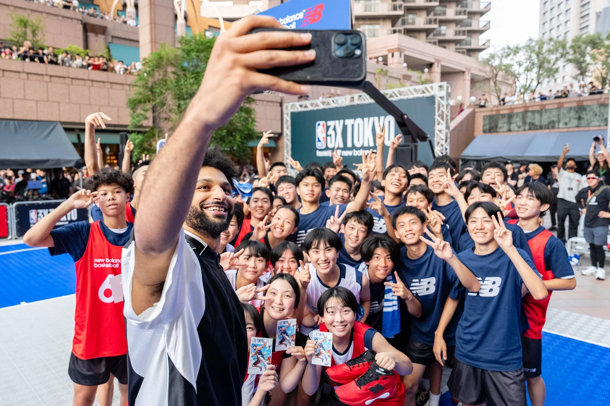 男性が多数の若者たちと屋外のバスケットボールイベントで自撮りしている様子。