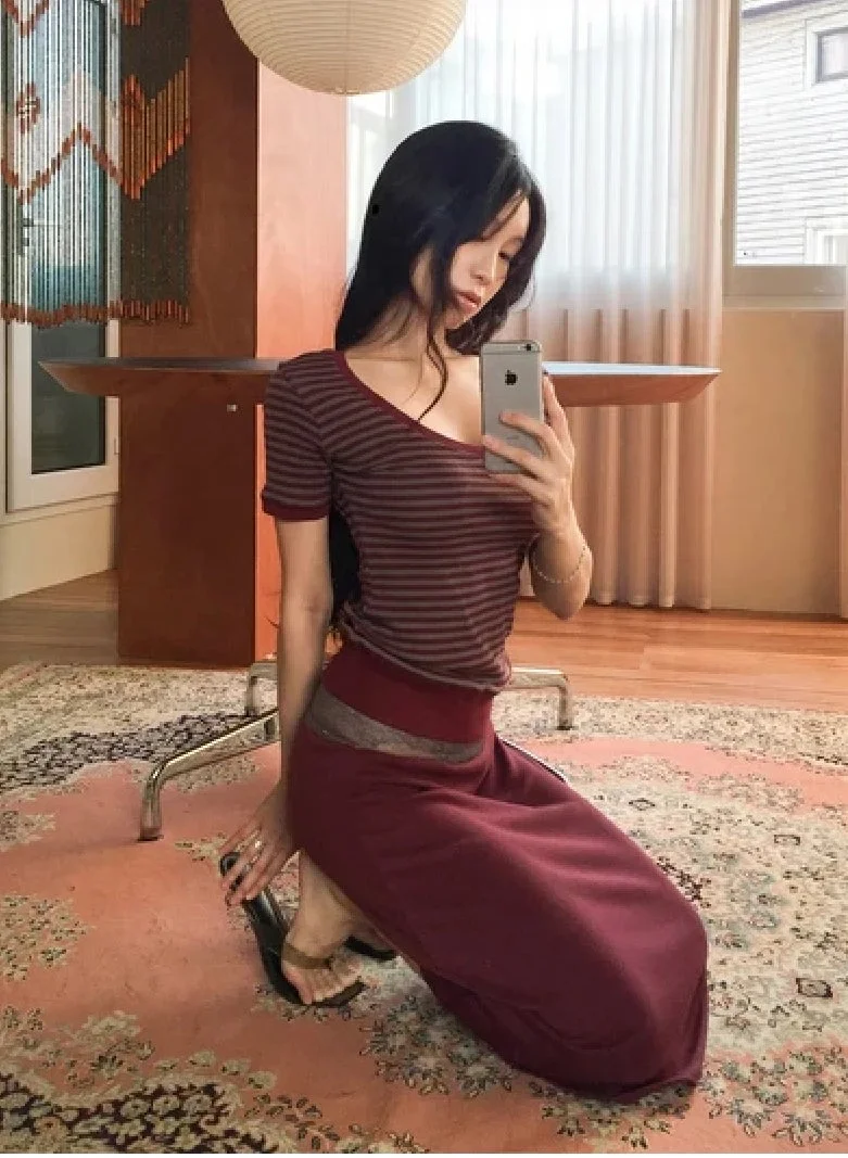 ストライプのトップスとワインレッドのロングスカートを着用した女性が、室内のカーペットの上でスマートフォンを持ち自撮りしている