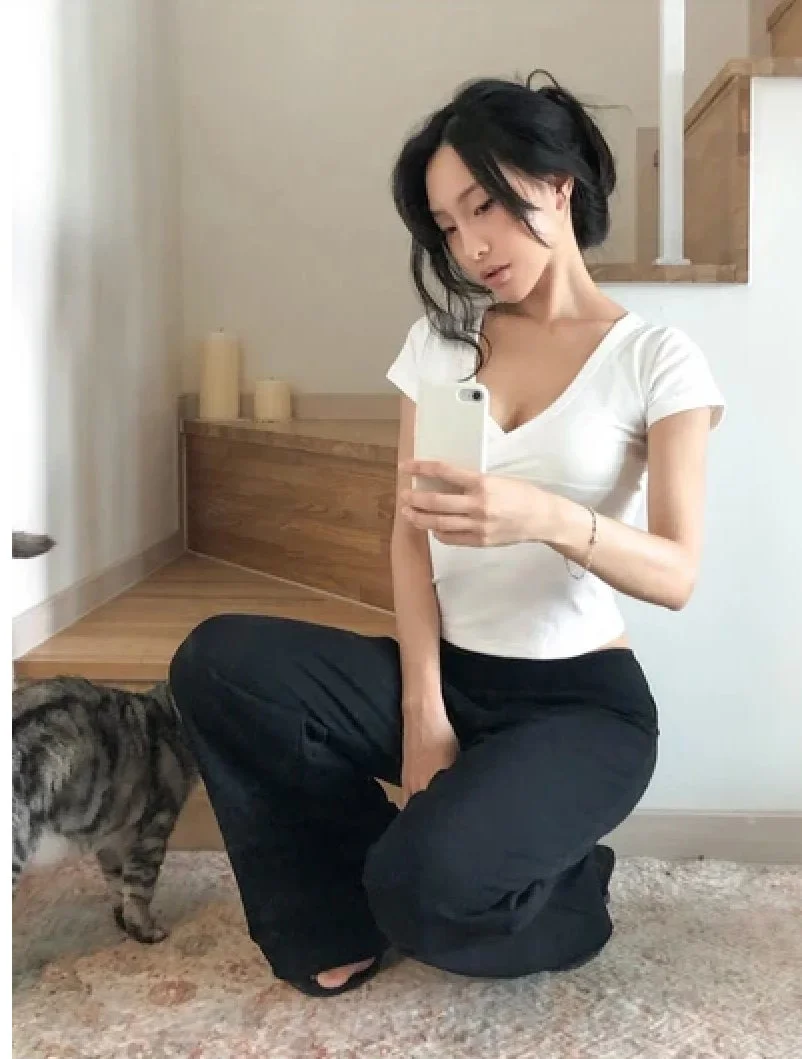 白いTシャツと黒いパンツ姿の女性が、室内の床にしゃがんでスマートフォンで自撮りしている