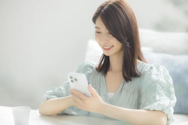 明るい室内で、笑顔の若い女性がスマートフォンを操作している。テーブルにはカップが置かれ、リラックスした日常の様子がうかがえる。