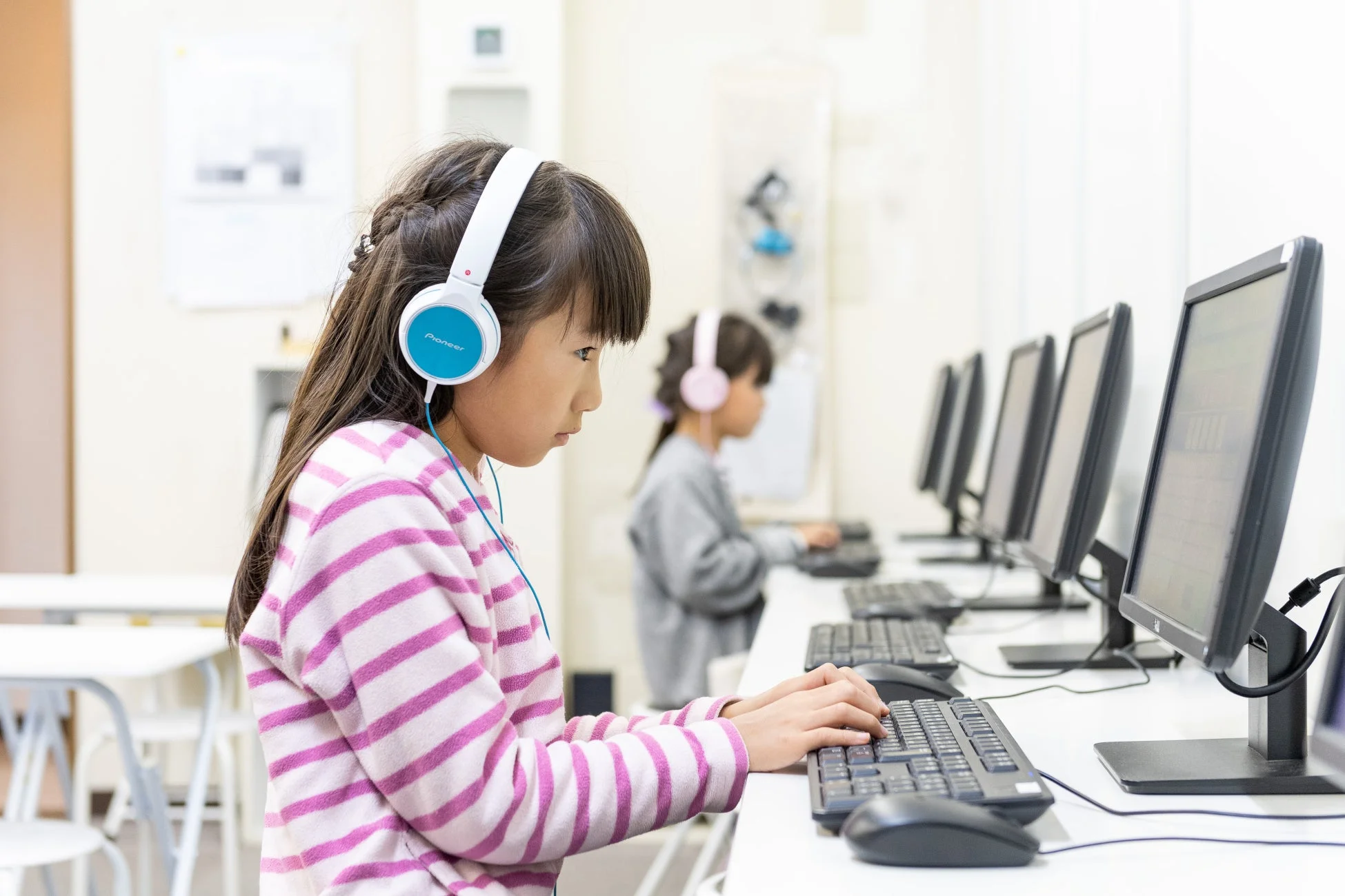 教室でヘッドホンを着用し、パソコンを使って学習する二人の子供たちの様子です。手前の女の子は真剣な表情でキーボードを打っています。