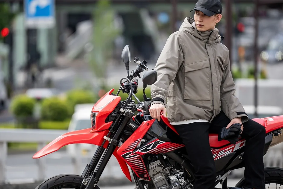ベージュのジャケットと黒いキャップを着用した男性が、赤いオフロードバイクに腰掛けている。