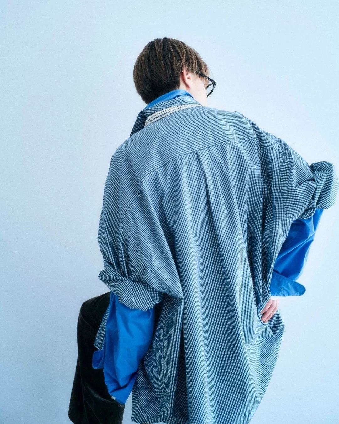 オーバーサイズのギンガムチェックシャツを着用し、明るいブルーのシャツとパールネックレスを合わせた人物の後ろ姿
