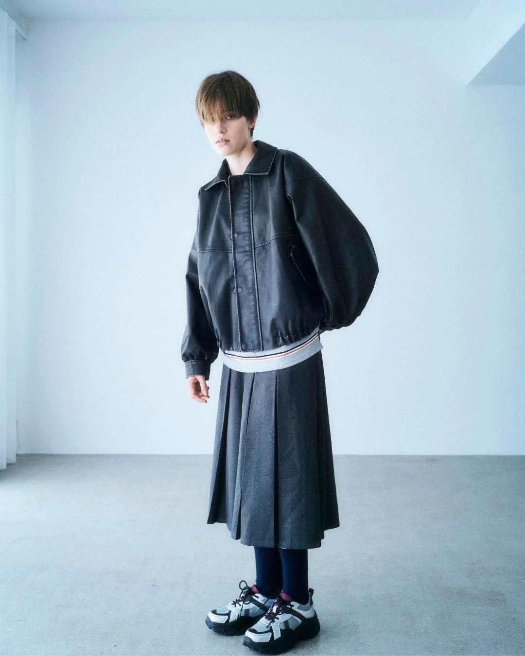 黒いブルゾンとグレーのプリーツスカート、厚底スニーカーを合わせたショートヘアの人物