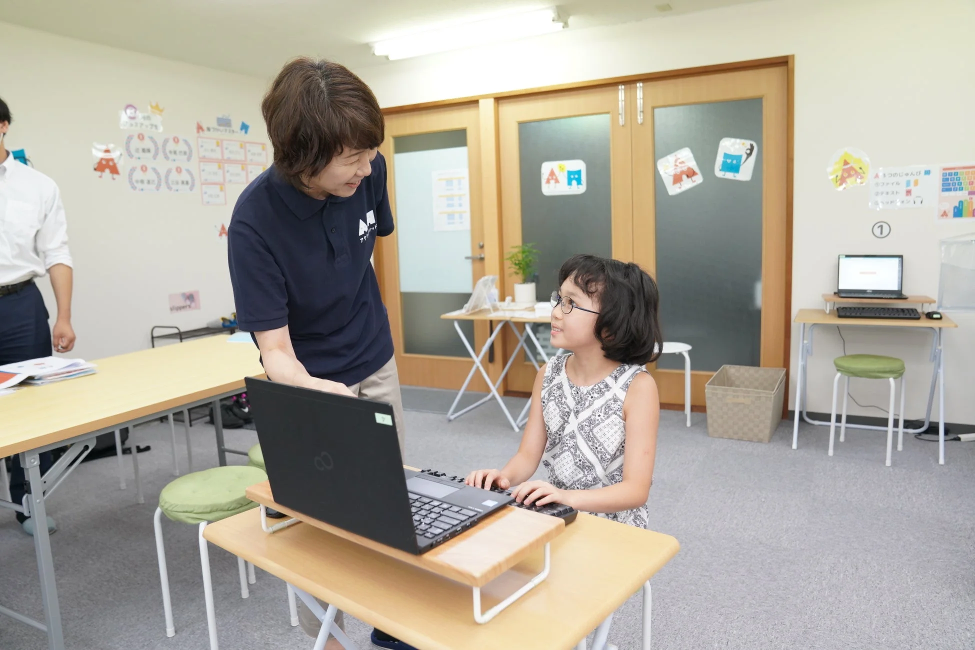 教室で女性の先生がノートパソコンを使用する女の子に個別指導を行っています。学習に集中する生徒と優しい眼差しの先生の姿から、和やかな雰囲気の中でデジタル学習が進められている様子が伝わります。